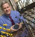 Mechanical engineer Kevin Hemker, seated between models representing how atoms are packed within an individual grain in a material, holds a silicon wafer onto which nanocrystalline aluminum thin film specimens have been deposited.