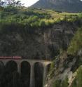 A train crosses a river gorge in the Swiss Alps that drops steeply from the floor of the broad glacial valley above it.