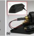 In transmission mode, the holographic microscope has a wider field of view than in reflection mode. While in this orientation, the device can be used to search for pathogens in large volumes of see-through materials, such as blood or water. (a) The beam cube, which splits the laser into two beams in reflection mode, is removed. Laser light from a laser diode (“LD”) is projected through a pin hole (“PH”) and passes directly through a see-through sample. Light that hits individual cells or particles in the sample is scattered; this scattered light forms an interference pattern with light that has not encountered any obstacles on its path through the sample. The interference pattern formed by the encounter between these two sources of light is then recorded on a CMOS image sensor and sent to a computer for analysis. (b) This photograph shows the microscope in transmission mode, with its cover removed. (The inset shows what the microscope looks like with its cover on.)