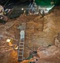 The excavation shows layered sequences of human prehistory captured in this cave. A yellow total station is shown at the right.